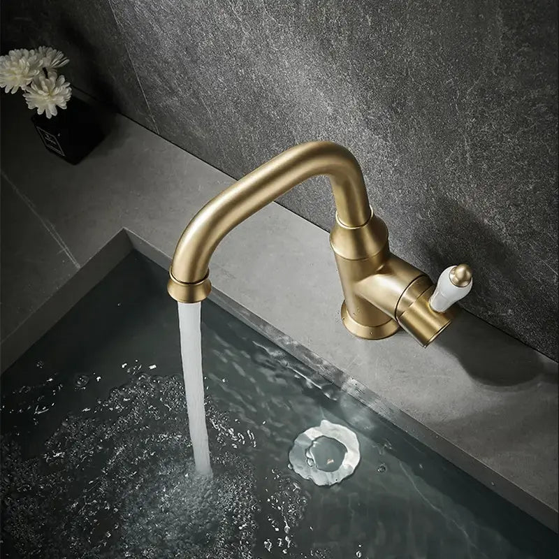 The Tiqui™ Single Handle Bathroom Sink Faucet, featuring a brass finish and white handle, is installed over a gray countertop, pouring a stream of water into the sink. The background features a dark textured wall and a small vase with white flowers.