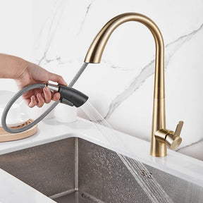 The Tiqui Single Lever Pull Out Spring Spout Faucet, featuring a sleek, curved design in brushed gold, is elegantly installed over a white sink. Water flows gracefully from the spout. The background showcases a white marble backsplash with subtle gray veining. Two white canisters with wooden lids sit on the counter, completing the sophisticated look.