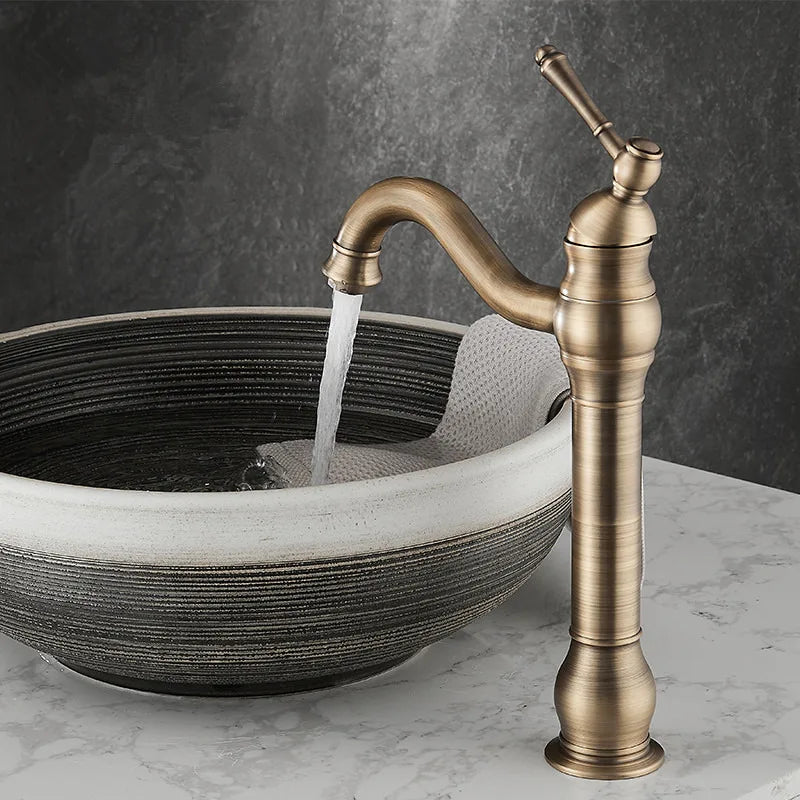 An Allfixture Deck Mounted Antique Brass Bathroom Faucet with an elegant curved spout and single handle stands beside a round, textured stone sink. Featuring a hot and cold water mixer, the tall, dark bronze faucet perfectly complements the folded beige towel draped over the sink edge against a dark gray marble wall and white marble countertop.