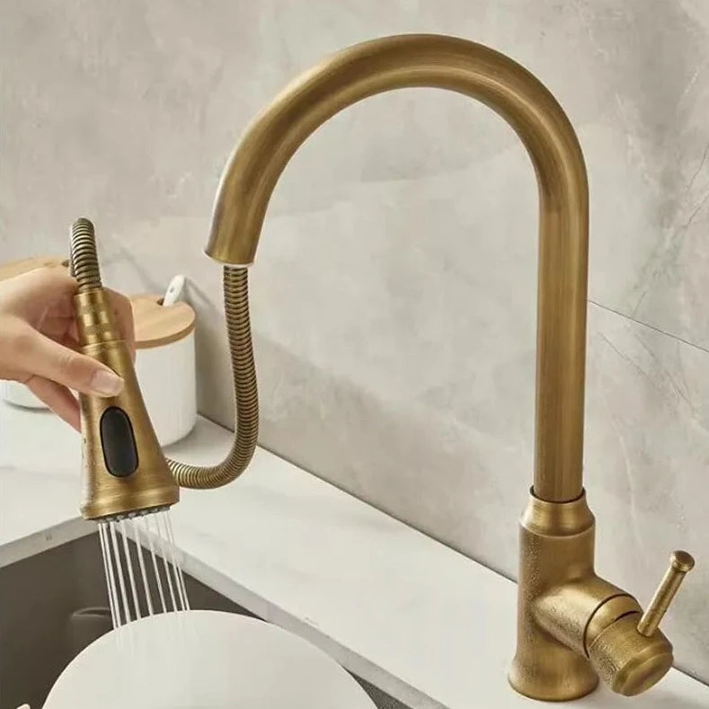 An Antique Brass Pull-Down Spout Kitchen Faucet by Rozin with a high arc and sleek, modern design pours water into a stainless-steel sink. The faucet handle is positioned to the right. In the background, reminiscent of elegant bathroom faucets, two white canisters with wooden lids are on a marble countertop.