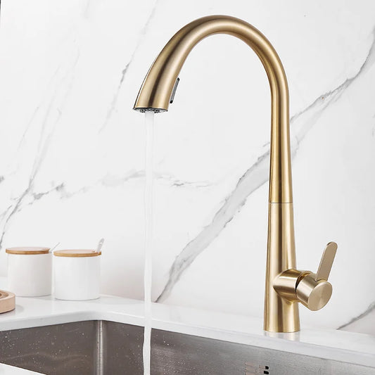 The Tiqui Single Lever Pull Out Spring Spout Faucet, featuring a sleek, curved design in brushed gold, is elegantly installed over a white sink. Water flows gracefully from the spout. The background showcases a white marble backsplash with subtle gray veining. Two white canisters with wooden lids sit on the counter, completing the sophisticated look.