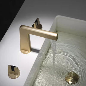 A sleek, modern Solid Brass Dual Handle Bathroom Faucet by Tiqui, featuring a rectangular spout in a brass finish, cascades water into a white sink. Flanked by two matching handles on each side, the minimalist design stands out against a dark gray backdrop. Complementing this piece are equally elegant Tiqui shower faucets to maintain a cohesive aesthetic.