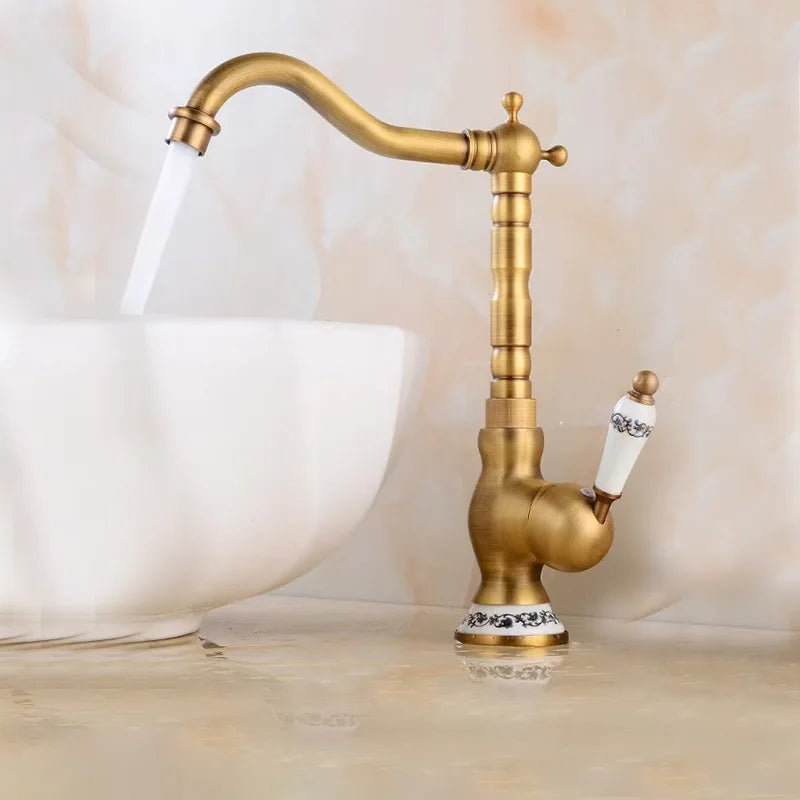 An Allfixture Tall Torneiras Bathroom Faucet – Single Handle, Hot and Cold Water Mixer, featuring a vintage-style brass finish with a white ceramic handle and decorative base, is installed next to a modern sink basin. Water flows from the faucet into the sink, set against a light beige background.