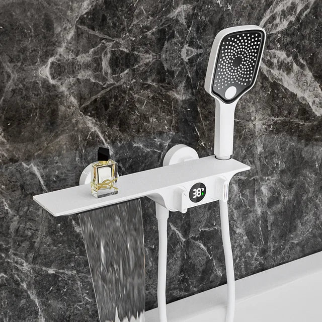 A modern shower setup features the Allfixture Wall-Mounted Digital Display Bath and Shower Faucet Set - Waterfall Shower Mixer, showcasing a sleek white showerhead with an attached rectangular shelf. The shelf holds a small bottle of liquid, while below it, water cascades from a waterfall spout. The backdrop is a dark, marbled wall, with the digital display faucet reading 38°C.