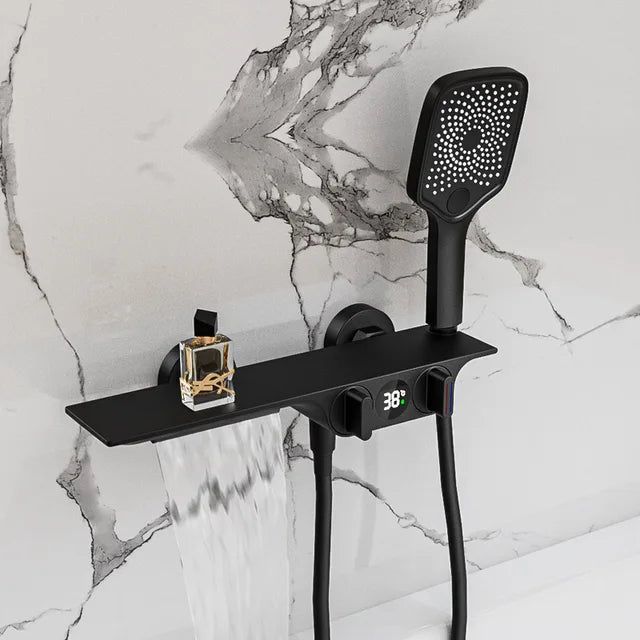 A modern shower setup features the Allfixture Wall-Mounted Digital Display Bath and Shower Faucet Set - Waterfall Shower Mixer, showcasing a sleek white showerhead with an attached rectangular shelf. The shelf holds a small bottle of liquid, while below it, water cascades from a waterfall spout. The backdrop is a dark, marbled wall, with the digital display faucet reading 38°C.