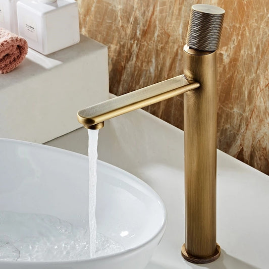 The Antique Bronze Bathroom Faucet by Tiqui features a sleek, cylindrical design in antique bronze. Water flows smoothly into a white, round basin sink. The stylish setup is complemented by a marble wall and a countertop adorned with various toiletries, making it one of the most elegant bathroom faucets available.