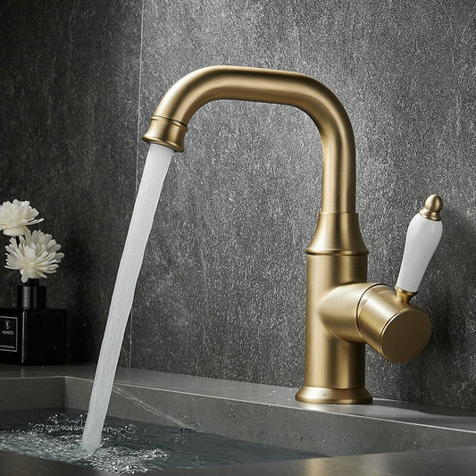 A Solid Brass Contemporary Single Handle Bathroom Faucet by Tiqui with a curved spout is installed over a gray stone sink. The faucet is turned on, and water flows from it. The handle of the faucet is white with a golden tip. A black soap dispenser and white flowers are visible in the background.