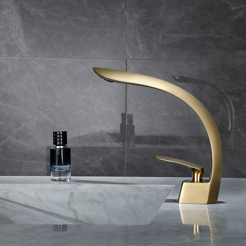 A sleek, modern Tiqui Wasser™ Single Handle Bathroom Sink Faucet with a curved spout and single lever handle is installed on a marble countertop. A small bottle of liquid soap or lotion sits nearby against a backdrop of gray tile walls.