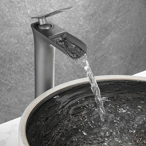 The Tiqui™ Gunmetal Single Handle Bathroom Sink Faucet, a tall and sleek stainless steel fixture with a modern design, dispenses water into a round, stylish stone sink set on a marble countertop. The background features a textured gray wall.