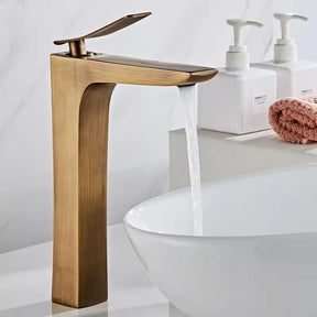 An Antique Bronze Deck Mounted Bathroom Sink Faucet by Tiqui is shown with water flowing from its spout into a white, round basin sink. The sleek design and minimalist aesthetics suggest a contemporary bathroom setting. The faucet handle is positioned upright.