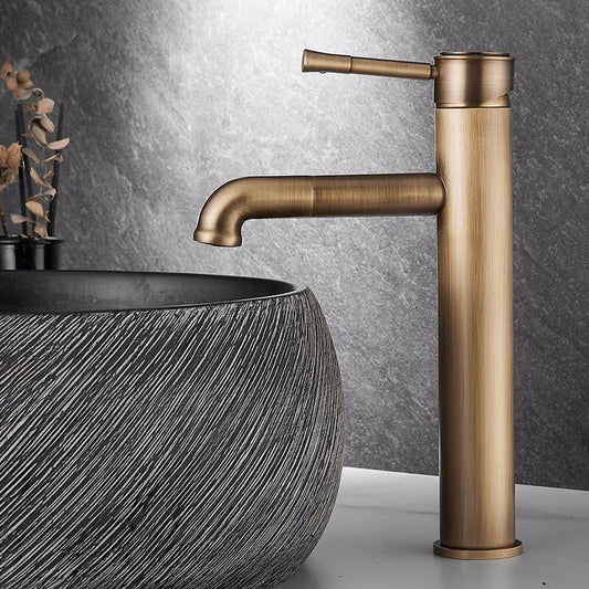 An Antique Bronze Single Handle Bathroom Sink Faucet from Tiqui stands tall beside a round, textured stone sink with a grey and white swirl pattern. Complementing the design is a sleek shower faucet in the background against the textured, stone-like grey wall, creating a stylish and minimalist bathroom aesthetic.