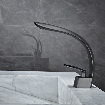 A sleek, modern Tiqui Wasser™ Single Handle Bathroom Sink Faucet with a curved spout and single lever handle is installed on a marble countertop. A small bottle of liquid soap or lotion sits nearby against a backdrop of gray tile walls.