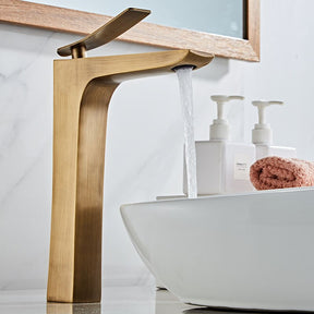 An Antique Bronze Deck Mounted Bathroom Sink Faucet by Tiqui is shown with water flowing from its spout into a white, round basin sink. The sleek design and minimalist aesthetics suggest a contemporary bathroom setting. The faucet handle is positioned upright.