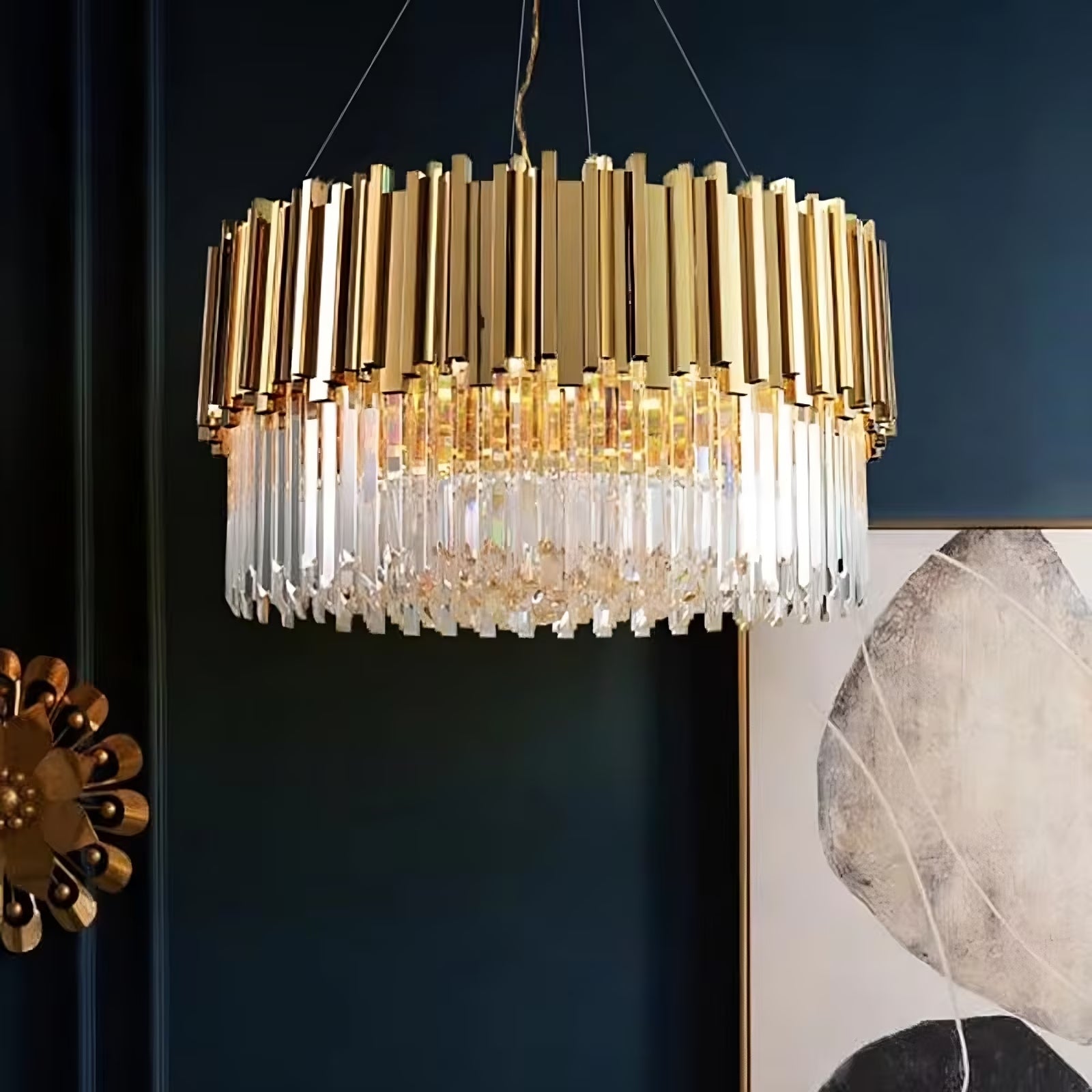 A luxurious Gio Gold Plated Crystal Chandelier from Morsale.com, featuring a combination of gold metal bars and hanging K9 crystal prisms, is suspended from the ceiling. The backdrop includes a dark wall with a large abstract painting in gray and white tones and a decorative gold wall piece.
