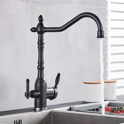 A sleek Uythner Dual Handle Solid Brass Faucet With Filtered Water Tap, featuring a vintage design, is mounted on a gray countertop. Water flows from the curved spout into a stainless steel sink below. In the background, a white bowl with a decorative pattern rests beside vegetables. Light filters through blinds, highlighting the elegant fixture.