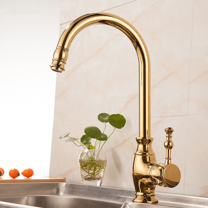 A Polished Gold Single Handle Kitchen Faucet by Tiqui with a curved spout is installed over a stainless steel sink. The background features a light-colored tiled wall, reminiscent of those you'd find in a bathroom with elegant bathroom sink faucets, along with a small glass vase holding a green plant and several small red tomatoes on the countertop.