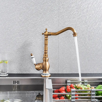 An Ellen Single Handle Antique Brass Kitchen Faucet, featuring a vintage-style design with a white ceramic handle and blue floral accents, is mounted on a marble countertop next to a sink filled with tomatoes and green peppers. A glass of water with lemon slices completes the charming setting.