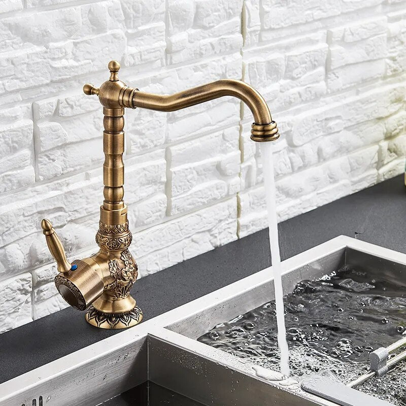 An Antique Brass Single Handle Faucet by Rozin, with its intricate carvings, streams water into a modern stainless steel sink against a textured white tile backsplash.