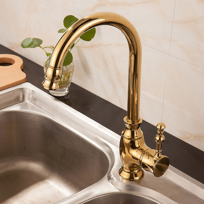 A Polished Gold Single Handle Kitchen Faucet by Tiqui with a curved spout is installed over a stainless steel sink. The background features a light-colored tiled wall, reminiscent of those you'd find in a bathroom with elegant bathroom sink faucets, along with a small glass vase holding a green plant and several small red tomatoes on the countertop.