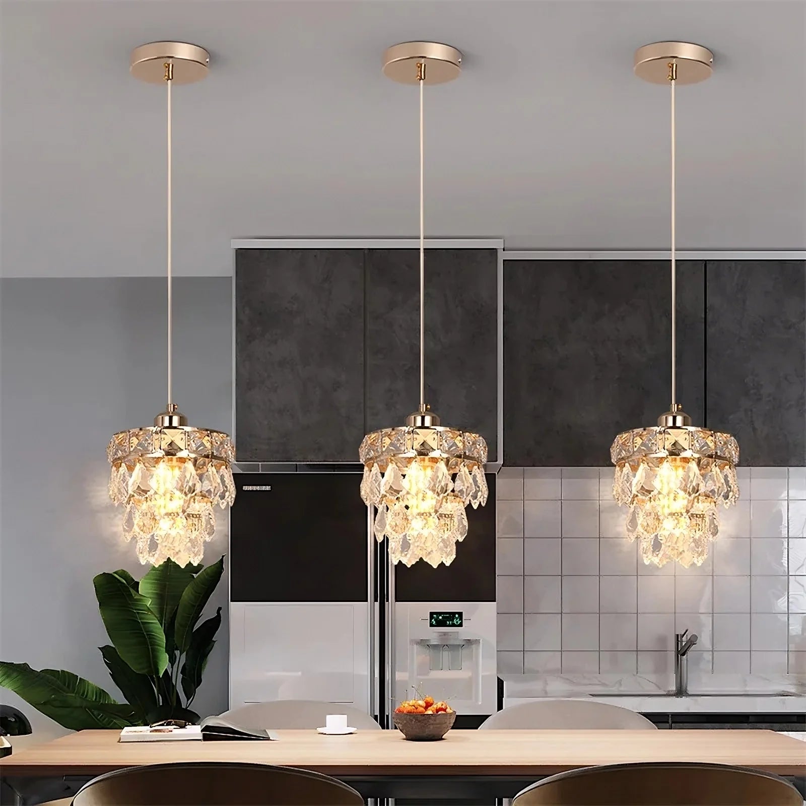 A modern kitchen features three Lazzo Kitchen Table Pendant Light Fixtures from Morsale.com with cascading handmade crystal accents hanging above a dining table. The background includes dark and light grey cabinetry, a refrigerator, and a tiled backsplash. A lush green plant adds a touch of nature to the scene.