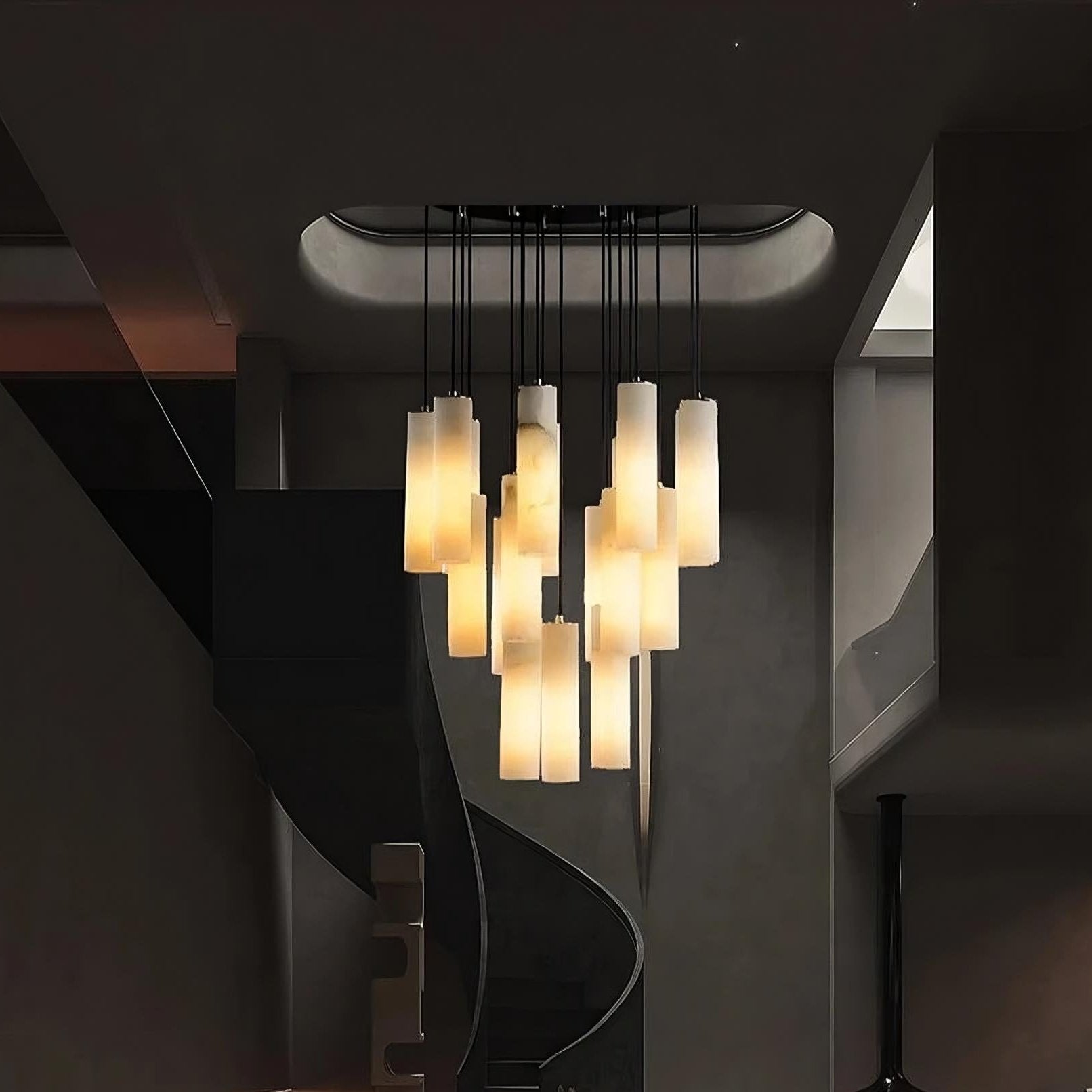 A modern chandelier with multiple cylindrical light fixtures hangs from the ceiling in the center of the room. The chandelier, featuring Morsale.com's Moonshade™ Marble Pendant Lighting Fixture, emits a warm glow, illuminating a winding black spiral staircase beneath it. The room has a minimalist design with dark and neutral tones.