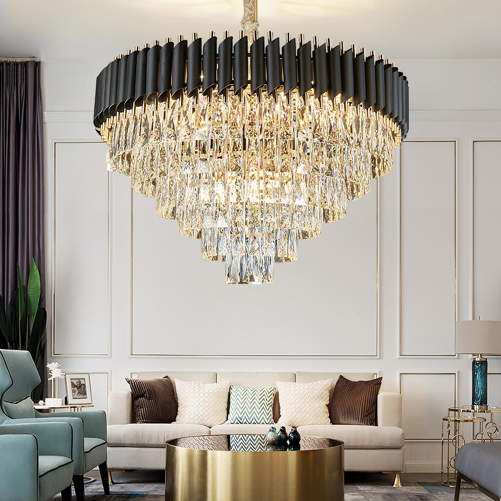 A luxurious, modern living room featuring the Verano Crystal Chandelier by Morsale.com with high-quality clear crystals in the center. The room boasts a beige sofa adorned with decorative cushions, a round gold coffee table, and elegant armchairs. The walls are beautifully paneled, and a small plant adds a touch of greenery in the corner.