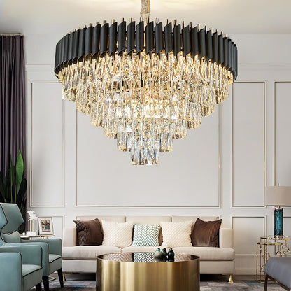 A luxurious, modern living room featuring the Verano Crystal Chandelier by Morsale.com with high-quality clear crystals in the center. The room boasts a beige sofa adorned with decorative cushions, a round gold coffee table, and elegant armchairs. The walls are beautifully paneled, and a small plant adds a touch of greenery in the corner.