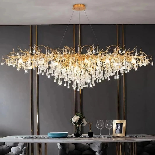 In the sophisticated dining room, the 62" Albero Dining Room Crystal Chandelier by Morsale.com, featuring intricate gold branches and hanging crystal droplets, is gracefully suspended above a modern dining table. The table is elegantly set with blue plates, a floral centerpiece, two wine glasses, and a framed picture. Dark walls serve as the background.