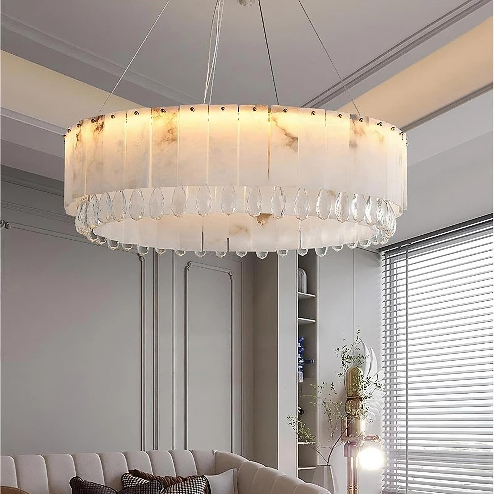 This modern living room, a luxury home decor highlight, features the Marble & Crystal Modern Chandelier by Morsale.com. The chandelier is large and circular with a frosted glass design. The room also boasts a light gray ceiling with crown molding, a tufted gray couch adorned with white pillows, and a floor lamp beside it. Blinds cover the window.