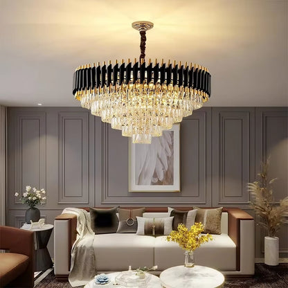 A luxurious living room features the Verano Crystal Chandelier from Morsale.com, showcasing cascading elements that epitomize modern design and create warm ambient lighting. The room includes a white sofa adorned with neutral cushions, a throw blanket, vases of flowers, and a round marble coffee table.