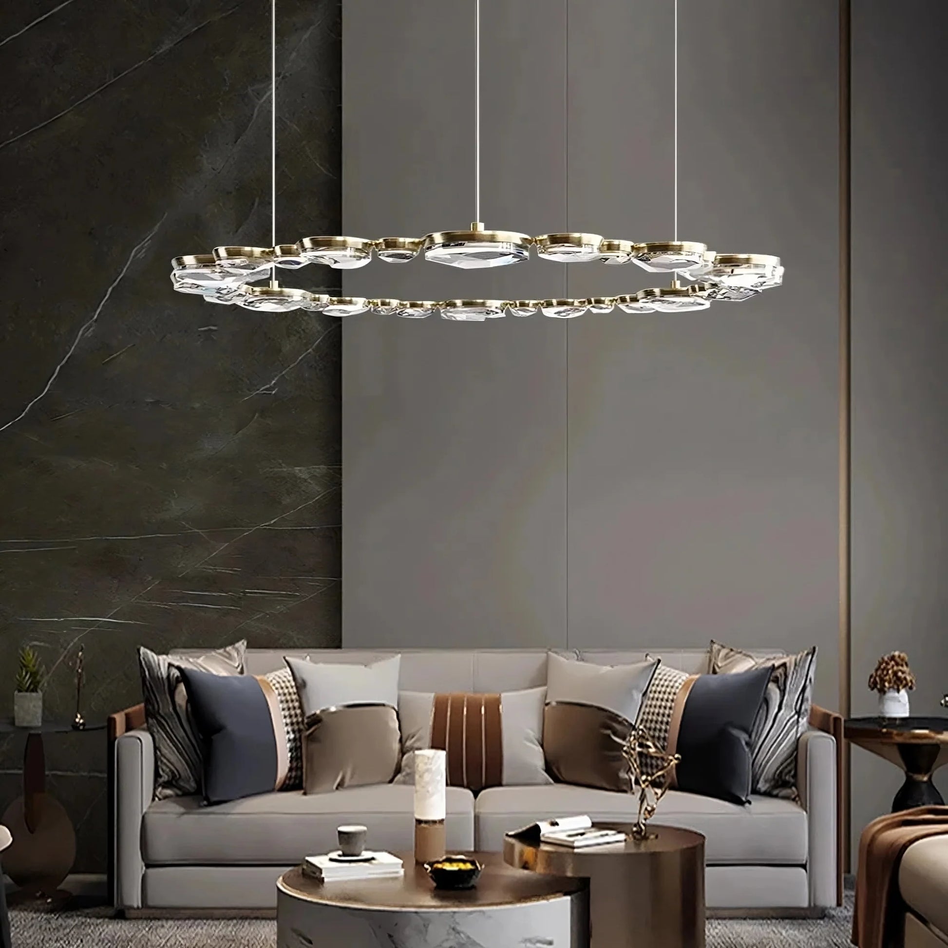 A modern living room showcases the Diamante Crystal Ceiling Light Fixture by Morsale, with glass and gold accents. Below is a gray sofa with patterned pillows, a round coffee table holding decorative items, and beige walls set off by a dark marble-like accent wall.