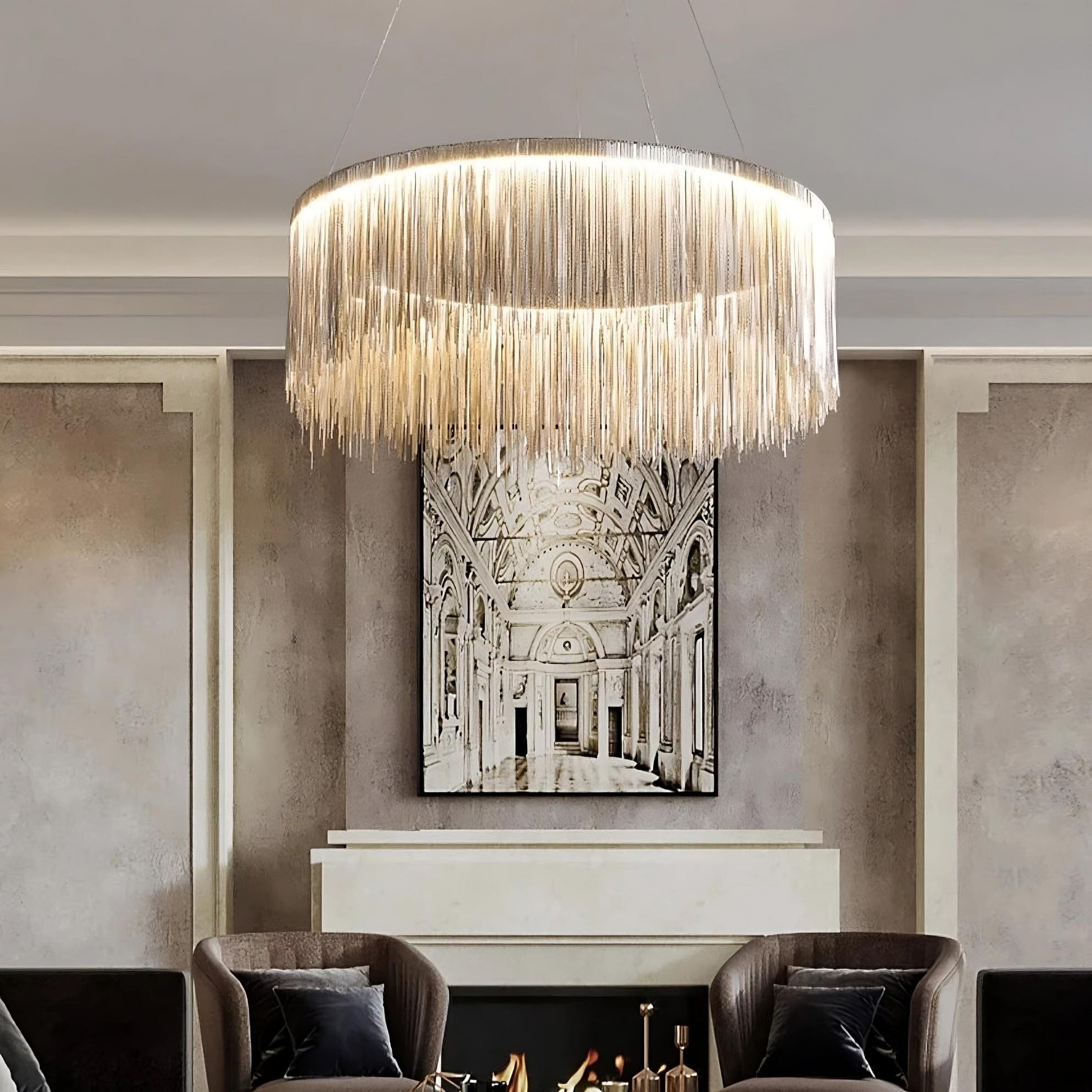 A modern living room features a Morsale.com Cadena Steel Tassel Light Fixture with cascading fringe hanging from the ceiling. Below, energy-efficient LED lighting highlights a fireplace with a black and white architectural painting mounted above it. Two dark armchairs with gray pillows are placed on either side of the fireplace.
