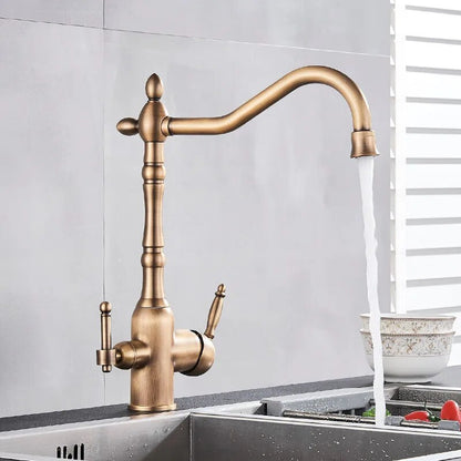 A sleek Uythner Dual Handle Solid Brass Faucet With Filtered Water Tap, featuring a vintage design, is mounted on a gray countertop. Water flows from the curved spout into a stainless steel sink below. In the background, a white bowl with a decorative pattern rests beside vegetables. Light filters through blinds, highlighting the elegant fixture.