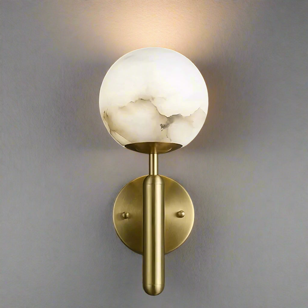A wall-mounted brass light source featuring a cylindrical base and a spherical, translucent white glass shade with marble-like veining. The background is a smooth, dark gray surface, highlighting the warm, elegant design of the dimmable Natural Marble Sphere Wall Sconce by Morsale.com.