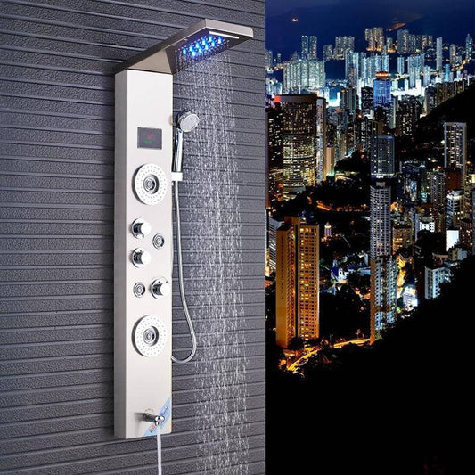 A **5-Function LED Shower Panel With Massage Jets Waterfall Rainfall** from Morsale.com, featuring multiple jets and a handheld showerhead, is mounted on a dark textured wall. The panel includes LED lighting and digital controls. Positioned near a sleek bathroom sink faucet, the scene reveals a cityscape with numerous high-rise buildings visible at night.