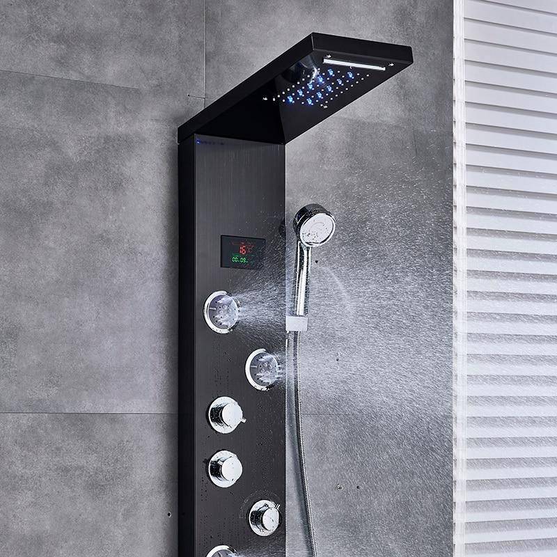 A Morsale.com 5-Stage LED Full Body Shower Panel With Massage Jets - Charcoal is mounted on a gray tiled wall. Water streams from the large overhead shower head, while multiple jets and a handheld showerhead enhance the modern design. In the background, there's a white bathtub with a towel draped over its edge.