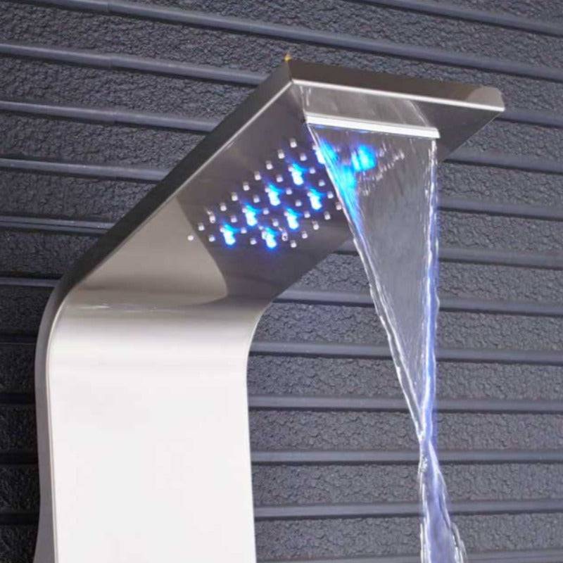 A woman in a white shirt stands in front of the Morsale.com 6-Function Wall Shower Panel with Massage Jets, Brushed Nickel, which features multiple controls and a rainfall showerhead spraying water in a bright, spacious bathroom. The shower faucet is illuminated by LED lights around the showerhead.