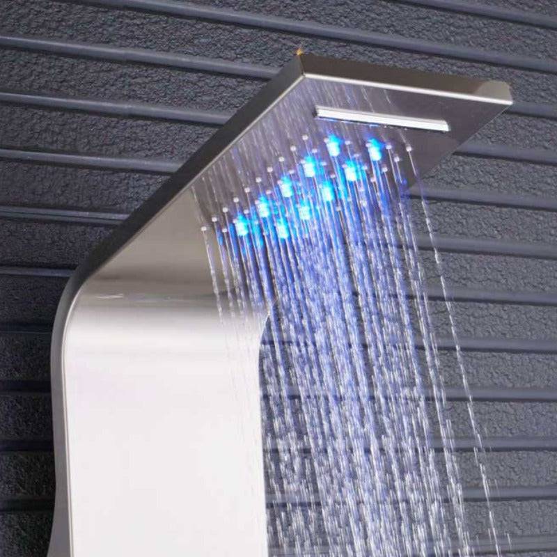 A woman in a white shirt stands in front of the Morsale.com 6-Function Wall Shower Panel with Massage Jets, Brushed Nickel, which features multiple controls and a rainfall showerhead spraying water in a bright, spacious bathroom. The shower faucet is illuminated by LED lights around the showerhead.