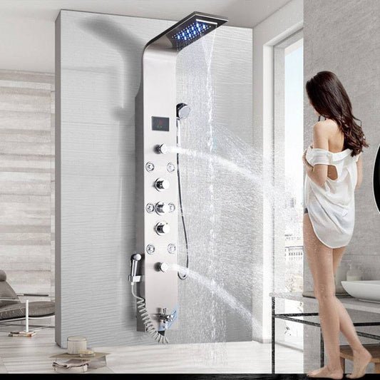 A woman in a white shirt stands in front of the Morsale.com 6-Function Wall Shower Panel with Massage Jets, Brushed Nickel, which features multiple controls and a rainfall showerhead spraying water in a bright, spacious bathroom. The shower faucet is illuminated by LED lights around the showerhead.