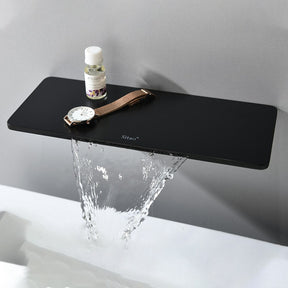 A modern bathroom sink with a minimalist design. The rectangular white sink has water pouring into it from the sleek, black Solid Brass Wall Mounted Waterfall Sink Faucet by AllFixture, which includes a stylish black handle. A shelf above the faucet holds a small container and a loofah. Towels are placed on the counter.