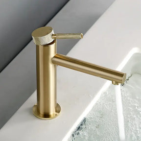 The allfixture Solid Brass Single Handle Bathroom Sink Faucet, featuring a modern gold-toned finish, cylindrical base, and sleek side-mounted handle, is shown. Water flows steadily from the spout into a white basin filled with water, creating ripples. The background is plain and neutral.