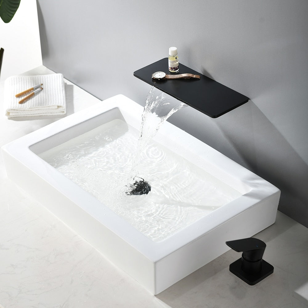 A modern bathroom sink setup featuring the AllFixture Solid Brass Wall Mounted Waterfall Sink Faucet extending horizontally from the wall in a gold finish. Water flows gracefully from the faucet into a rectangular white basin. Two small bottles are placed on the faucet shelf above the cascading water. The wall and countertop are decorated in muted tones.