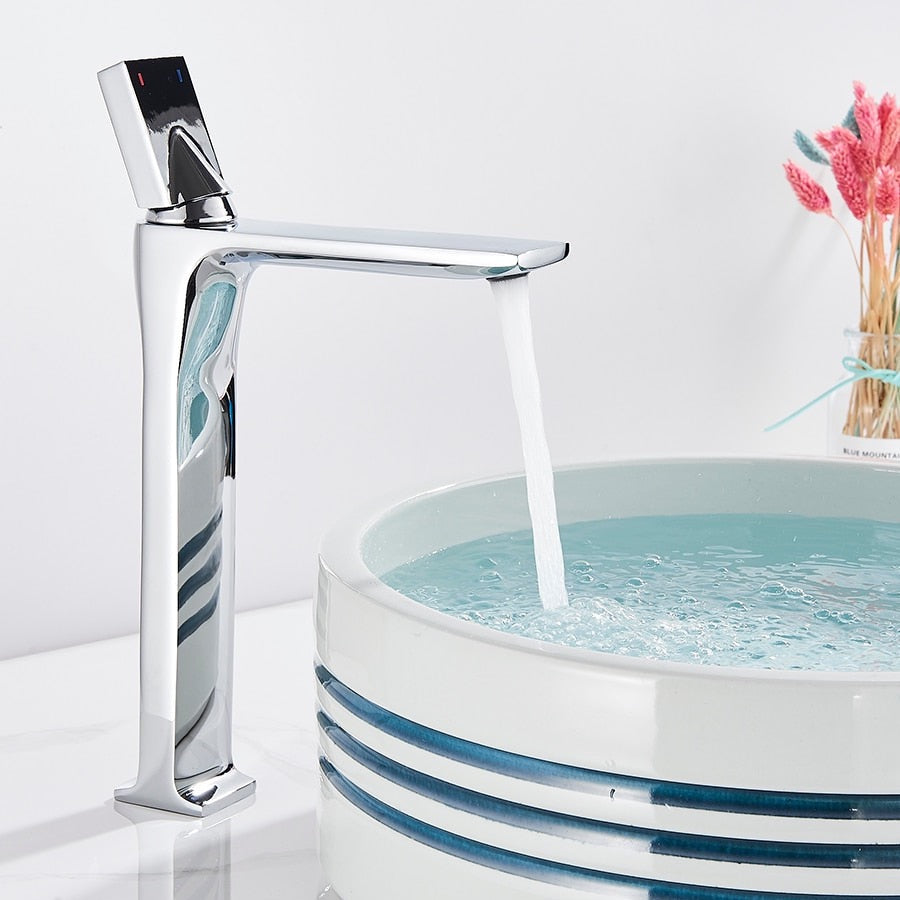 An AllFixture Deck Mounted Brass Single Handle Brass Basin Faucet in modern black matte is attached to a white counter. It is pouring water into a white, round basin with blue stripes near the rim. The counter is decorated with a vase of colorful dried flowers in the background.