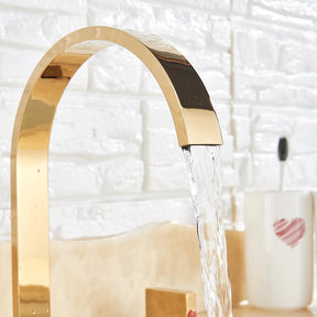 A modern bathroom sink features the Double Handle Deck Mounted Bathroom Faucet by AllFixture in polished gold, with a high-arching spout that gracefully dispenses water into the basin. The faucet has two square-shaped handles; one bears a red indicator for hot water, and the other displays a blue indicator for cold water. In the background, a round mirror can be seen.