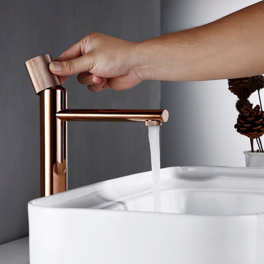 A sleek, polished rose gold bathroom faucet from Smesiteli with a contemporary design dispenses water into a modern, white rectangular sink, which is partially filled with water. The sink is positioned on a white bathroom countertop. A small decorative item is visible in the background.