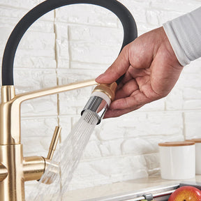 A modern kitchen sink with the sleek, gold-toned Solid Brass Kitchen Faucet With Filtered Water Tap by allfixture, featuring a black curved spout. The faucet is turned on, with water flowing over fresh produce in a stainless steel sink. In the background, there's a white brick wall and partially open window blinds.