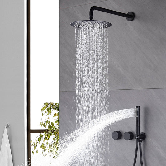 The Aodei Wasser™ Complete Shower System in Brushed Black showcases a modern setup, featuring water flowing from a large, round overhead showerhead. The system includes a detachable handheld shower sprayer that sprays water horizontally. The wall behind is tastefully tiled and accented by sleek bathroom faucets, with a small potted plant visible in the background.