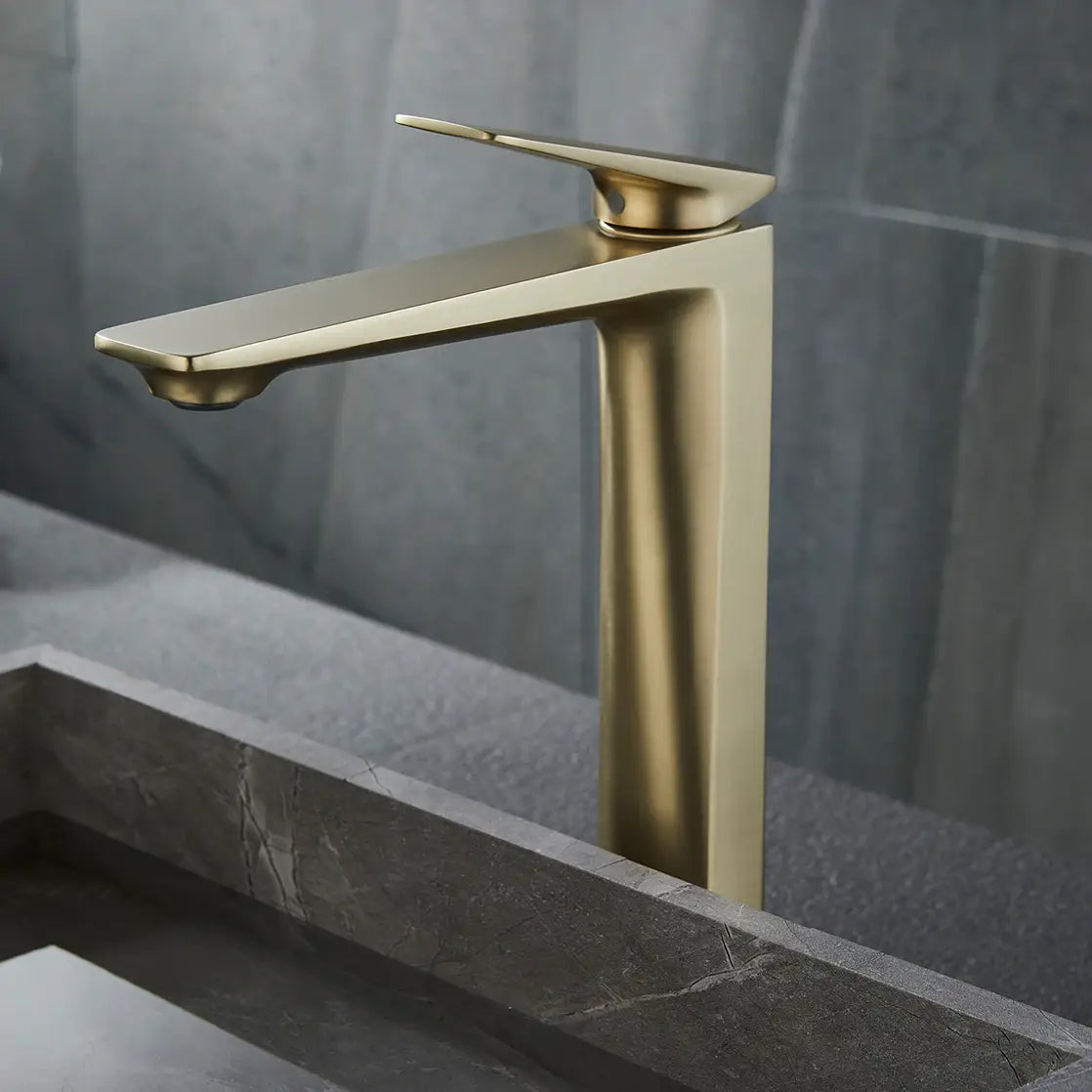 A modern, gold-colored Bagnolux Single Handle Brass Bathroom Sink Faucet with a sleek design is mounted on a grey stone countertop, pouring water into a matching rectangular grey stone sink. The backdrop features a textured grey wall, completing this elegant and cohesive look.