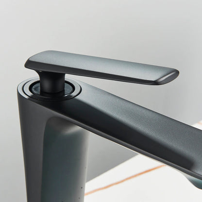 A Wasser™ Solid Brass Bathroom Sink Faucet by Rozin, featuring a sleek, angular design in modern black, dispenses water into a round, white vessel sink. In the background, a partial reflection is seen in a circular mirror, while a small plant adds a touch of greenery to the scene.
