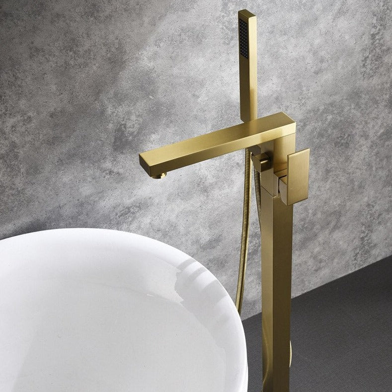 A modern brass Wasser™ Floor Mounted Bathtub Filler Shower Set by allfixture is installed against a gray wall. Water sprays horizontally from the handheld shower head attached to the fixture. The sleek, angular design and lever handle for controlling flow enhance its elegance. Below, the edge of a white bathtub is visible.